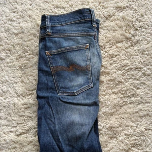 Blå jeans från Nudie Jeans, W27 L30 - Snygga blå jeans från Nudie Jeans med klassisk femficksdesign och coola orangea sömmar. Jeansen har en rak passform och är tillverkade i slitstarkt denim med Nudie Jeans logga på bakfickan. Perfekt för dig som gillar stilrena och tidlösa jeans.