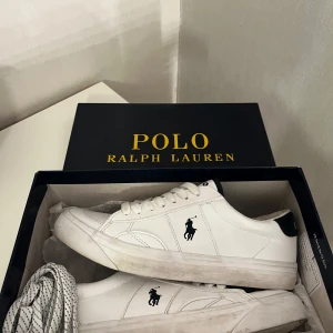 Vita sneakers från Polo Ralph Lauren -  Vita sneakers från Polo Ralph Lauren med svart logga på sidan och svart detalj vid hälen. Fint skick, använda ett få tal gånger. Klassisk design med Polo-tryck på plösen och insidan. Ingår extra snören och original box. Pris går att diskutera.