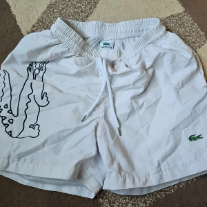 Vita badshorts från Lacoste - Snygga vita badshorts från Lacoste. De e sköna att ha på. Den är använd några gånger och är i bra skick.
