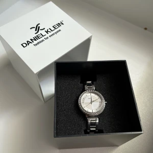 Silverfärgad klocka från Daniel Klein - Stilren och rund klocka från Daniel Klein i silverfärgat metall med länkarmband. Urtavlan är ljus med diskreta markeringar och ramen är dekorerad med glittrande stenar för extra bling. Perfekt accessoar för dig som gillar snygga detaljer.