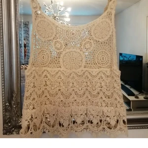 Beige virkad linnetopp med spetsdetaljer - Superfin beige linnetopp med breda axelband och genomgående virkad spets. Toppen har ett unikt cirkelmönster upptill och vågiga spetskanter nedtill som ger boho-vibbar. Perfekt att styla över en enkel topp eller bikini för en chill look.