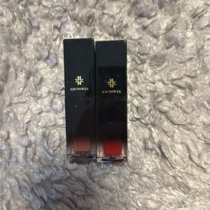 Lip Power läppstift Tiphaine & Philou - Två Lip Power läppstift i nyanserna Tiphaine (nude/brun) och Philou (röd). Snygga fyrkantiga svarta hylsor med guldtext. Perfekt för dig som vill ha både en neutral och en djärv färg till läpparna. Båda har flytande formula och är enkla att applicera.