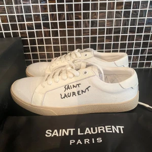Saint Laurent vita sneakers i canvas - Snygga vita sneakers från Saint Laurent med handskriven logga på sidan. Skorna har klassisk låg profil, rund tå och platt sula i beige gummi. Ovandelen är i canvas med vita snören och diskreta sömmar. Perfekt för dig som gillar stilrena och exklusiva sneakers.