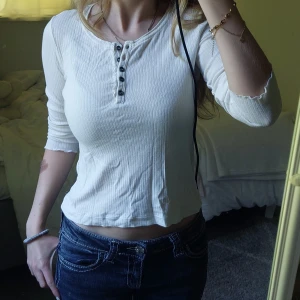 Vit ribbad topp med knappar - Säljer en vit ribbad långärmad tröja från Lindex i storlek xxs. Toppen har en rund halsringning och fem svarta knappar framtill. Materialet är mjukt bomullstyg och modellen är slim. 