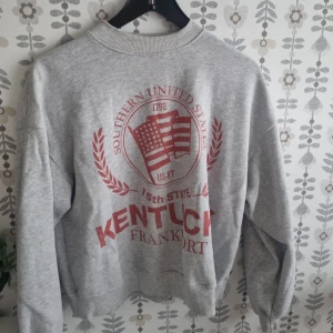 Grå sweatshirt Kentucky print - Säljer en grå sweatshirt med rött Kentucky-tryck och amerikansk flagga på framsidan. Tröjan har rund hals, ribbade muddar och lång ärm. Perfekt för dig som gillar college-stil och vill ha en chill look.