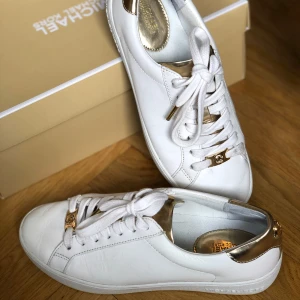 Vita sneakers från Michael Kors - Snygga vita sneakers från Michael Kors med detaljer i guld på hälen och vid snörningen. Skorna har rund tå, platt sula och är tillverkade i skinn. Perfekta för dig som gillar stilrena och lyxiga sneakers med en touch av glamour.
