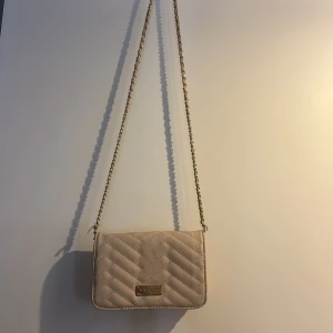 Beige quiltad axelväska från Bebe - Snygg beige axelväska från Bebe med quiltad design och guldfärgad kedja. Väskan har ett metallmärke framtill, flera fack inuti och en dragkedja för småsaker. Perfekt storlek för det viktigaste och ger en clean look till din outfit.