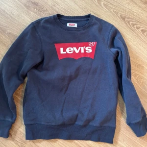 Mörkblå sweatshirt från Levi's - Säljer en mörkblå sweatshirt från Levi's med den klassiska röda loggan tryckt på bröstet. Tröjan har rund hals, långa ärmar och ribbade muddar vid ärmslut och nederkant. Perfekt för dig som gillar streetstyle och ikoniska märken.