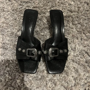 Svarta klackade sandaler med nitar - Svarta sandaler från H&M med låg klack och fyrkantig tå. Skorna har snygga silverfärgade  nitar som ger en cool vibe. Tillverkade i skinnimitation med platt sula framtill och öppen häl. Perfekta för dig som gillar statement-detaljer.