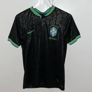 Svart Brasilien fotbollströja Nike - Snygg svart Brasilien fotbollströja från Nike med subtilt leopardmönster i tyget. Tröjan har gröna och blå detaljer på krage och ärmslut, samt Brasilien-emblem och Nike-logga på bröstet. Tillverkad i lätt, glansigt funktionsmaterial som andas.