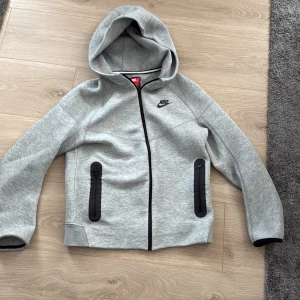 Grå tech fleece Nike - Snygg grå tech fleece från Nike med svart logga på bröstet. Jackan har hel dragkedja framtill, två svarta zip-fickor och huva. Materialet är mjukt och känns sportigt, perfekt för chill eller träning. Svarta detaljer vid ärmslut och fickor ger en clean look.  Änvänd en gång 