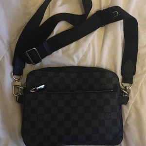 Louis Vuitton rutig axelväska svart - Säljer en snygg axelväska från Louis Vuitton med klassiskt rutigt mönster i svart och grått. Väskan har justerbar axelrem med Louis Vuitton-logga, silverfärgade metalldetaljer och dragkedja upptill. Perfekt storlek för dina viktigaste grejer.