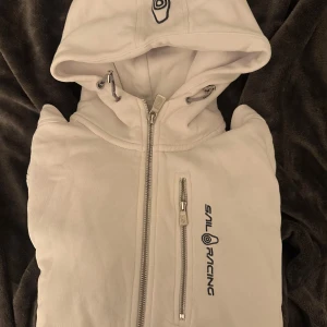 Vit hoodiejacka från Sail Racing - Snygg vit hoodiejacka från Sail Racing med dragkedja framtill och på bröstfickan. Jackan har broderad logga på bröstet och huvan, samt snörning i huvan för extra stil. Perfekt för dig som gillar sportig och clean look.