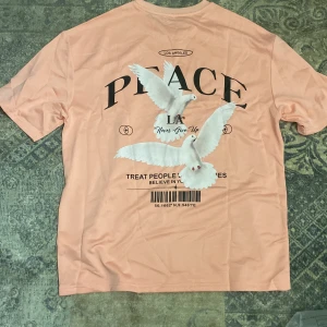 Persikofärgad t-shirt med tryck - Persikofärgad t-shirt med coolt tryck av vita duvor och texten 'PEACE' både fram och bak. T-shirten har rund hals och korta ärmar. Materialet känns mjukt och lätt, perfekt för chill dagar. Snygg grafisk design med LA-vibe och barcode-detalj på ryggen.