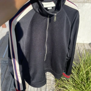 Svart Gucci zip - Gucci trackjacka i svart med vita och röda ränder längs ärmarna, samt broderad logga på ena ärmen. Jackan har dragkedja framtill och ribbade muddar med gröna och röda detaljer. Snygg sportig look och tillverkad i mjukt syntetmaterial.