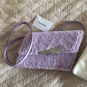 En helt ny ljuslila axelväska Acne Studios - Säljer en unik ljuslila axelväska från Acne Studios med fluffig frotté på utsidan och slät insida i matchande lila. Väskan har ett justerbart axelband och en snygg silverfärgad detalj med logga på framsidan. nypris: 6000kr