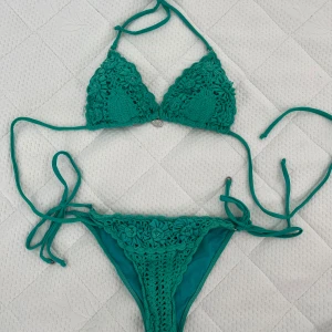 Turkos virkad bikini från RC - Snygg turkos bikini från RC med virkad design och blommiga detaljer på både överdel och underdel. Triangeltopp med knyt i nacken och ryggen, samt knyt på sidorna av trosan. Materialet är syntet med virkade inslag och pärlor i blommorna. Perfekt för stranden i sommar!