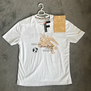 Vit Burberry t-shirt med tryck - Snygg vit t-shirt från Burberry med stort broderat logotryck i guld och vitt på bröstet. Klassisk rund hals med Burberrys rutiga detalj på insidan. Kortärmad och tillverkad i mjuk bomull. Det står st L fast den är mer xs/s