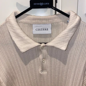 Beige ribbad stickad tröja från Ciszere - Säljer en beige stickad från Ciszere med ribbad struktur och klassisk krage. Tröjan har två knappar framtill och är tillverkad i ett mjukt stickat material som ger en chill och stilren vibe. Perfekt för dig som gillar enkel men snygg design.