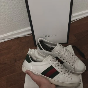 Gucci Ace sneakers - Säljer ett par klassiska Gucci Ace sneakers i vitt skinn med gröna och röda ränder på sidan. Skorna har snörning, rund tå och platt sula. Hälpartiet är grönt med textur och insidan har Gucci-logga. Perfekta för dig som gillar lyxiga detaljer och stilren design. 