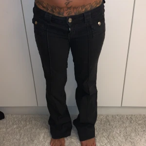 Svarta bootcut byxor med knappar - Snygga svarta byxor med bootcut passform och coola silverfärgade knappar fram. Byxorna har låg midja och markerade sömmar som ger en schysst look. Perfekta för dig som gillar en klassisk men ändå trendig stil. Materialet känns mjukt och bekvämt. Midjemått: 37cm