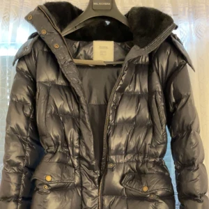 Svart pufferjacka från Boomerang XS - Svart pufferjacka från Boomerang i modellen ”Anna Down Jacket” i storlek XS. Jackan har en glansig yta, quiltad design och huva med mjukt foder. Praktiska fickor med knappar och dragkedja framtill. Perfekt för kalla dagar med sin varma och fluffiga känsla. Den är köpt för 8000kr förra året men jag säljer den för 1700kr. Kan eventuellt sänka priset vid snabb affär 🧥 