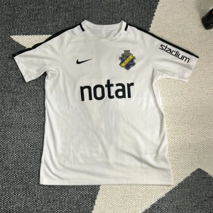 AIK vit matchtröja Nike  - AIK:s officiella vita matchtröja från Nike i barn XL, med svart tryck och detaljer. Tröjan har korta ärmar, Dri-FIT-material som andas, AIK:s klubbmärke på bröstet och sponsorloggor som Notar, Stadium, Fabege och SVEA. Snygg svart rand längs axlarna och nummer 17 på ryggen.