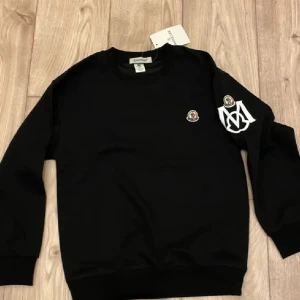 Svart sweatshirt från Moncler - Svart sweatshirt från Moncler med rund hals och långa ärmar. Tröjan har två broderade Moncler-loggor, en på bröstet och en på ärmen tillsammans med en vit M-detalj. Mjuk bomullskvalitet och ribbade muddar vid ärmslut och nederkant.