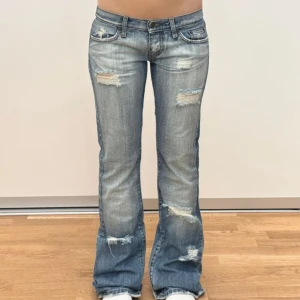 Blå bootcut jeans med slitningar - Snygga blå jeans med bootcut passform och slitna detaljer på både fram- och baksidan. Jeansen har låg midja, klassiska fem fickor och är tillverkade i jeansmaterial. Perfekt för dig som gillar en avslappnad och trendig stil med lite edge. Midja: 38 Innerben: 85