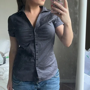 Mörkgrå kortärmad skjorta från H&M - Snygg mörkgrå skjorta från H&M med korta ärmar och figurnära passform. Skjortan har knappar hela vägen framtill och lätt rynkade detaljer vid bysten. Materialet är en mix av bomull, polyester och elastan som ger en skön stretch. Perfekt till jeans eller kjol!