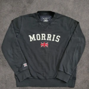 Svart sweatshirt från Morris med tryck - Svart sweatshirt från Morris i storlek medium med broderat vitt MORRIS-tryck och en röd Union Jack-flagga på bröstet. Tröjan har rund hals, ribbade muddar och är långärmad. Perfekt för dig som gillar klassisk brittisk stil och snygga detaljer.
