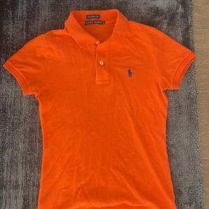 Orange polotopp från Ralph Lauren - Snygg orange polotopp från Ralph Lauren, modell The Skinny Polo. Klassisk pikékrage, två vita knappar och blå broderad logga på bröstet. Slim passform och korta ärmar, perfekt för dig som gillar färgstarka plagg med ikonisk stil.
