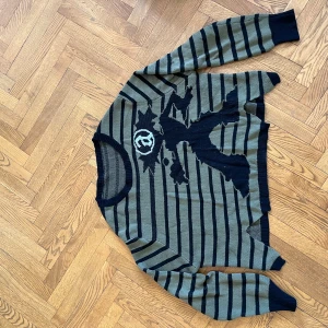 Randig stickad tröja med tryck - Cool stickad tröja från Acne Studios i olivgrönt och svart med randigt mönster och en stor grafisk figur på framsidan. Tröjan har rund hals, långa ärmar och ribbade muddar. Perfekt för dig som gillar streetstyle och unika prints.