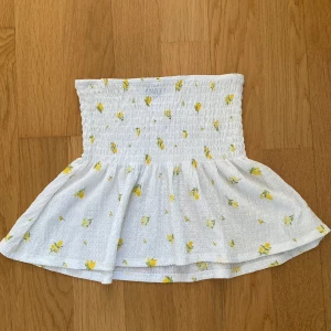 Vit blommig kortkjol från Gina Tricot - Supersöt vit kortkjol från Gina Tricot med smockad midja och utsvängd volang. Kjolen har små gula citroner och gröna blad som mönster, vilket ger en fräsch och somrig vibe. Perfekt för dig som gillar en feminin och lekfull stil.