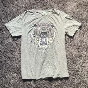 Grå Kenzo t-shirt med tigertryck - Storlek M-L Säljer en grå t-shirt från Kenzo med ikoniskt tigertryck och Kenzo Paris-logga på bröstet. T-shirten har rund hals, korta ärmar och är gjord i mjuk bomull. Perfekt för dig som gillar streetwear och vill sticka ut med ett coolt motiv.