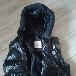 Svart dunväst från Moncler med huva - Svart glansig dunväst från Moncler med stor huva och klassisk logga på bröstet. Västen har en quiltad design och dragkedja framtill. Insidan har en cool serietidningsetikett och röda detaljer. Perfekt för att maxa din streetstyle under kyliga dagar.