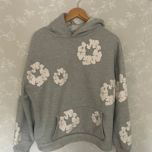 Grå hoodie med blommor från Denim Tears - Grå hoodie från Denim Tears med vita blommiga tryck över hela tröjan. Oversized passform, stor huva och klassisk magficka. Mjuk bomullsblandning som känns skön mot huden. Perfekt för dig som gillar streetwear med unik design.