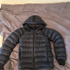 Peak Performance jacka - Säljer en svart dunjacka från Peak Performance med huva och dragkedja. Jackan är quiltad och har två fickor med dragkedja framtill. Perfekt för kalla dagar och har en clean, sportig look. Broderad logga på bröstet och elastiska muddar vid ärmslut.