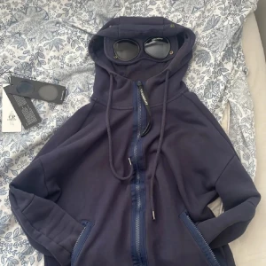 C.P. Company - Säljer en hoodie från C.P. Company med ikoniska goggles i huvan. Västen har dragkedja framtill, fickor med dragkedja och är gjord i mjuk bomull. Perfekt för dig som vill sticka ut med streetwear-vibe och coola detaljer. Skulle säga att den är 8/10 väldigt fin tröja skriv vid minsta fråga ☺️