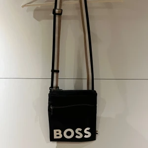 Svart axelväska från BOSS - Snygg svart axelväska från BOSS med justerbar rem i beige och svart. Väskan har en dragkedja framtill och stor vit BOSS-logga på framsidan. Perfekt storlek för det viktigaste och en clean look som passar till allt.