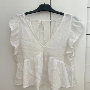 Vit volangblus med knyt i ryggen - Supersöt vit blus med volangärmar och v-ringning. Blusen har knyt i ryggen och är gjord i ett lätt, luftigt bomullstyg. Snygg på båda sommare och hösten med en kofta till. Hör av er vid frågor💖