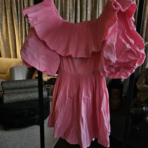 Rosa jumpsuit från H&M - Supersöt rosa jumpsuit med stora volangdetaljer och plisserad offshoulder-ringning. Klänningen har puffiga korta ärmar och en luftig, flowy passform som känns både trendig och lekfull. Perfekt för dig som vill sticka ut med en färgglad och feminin look.