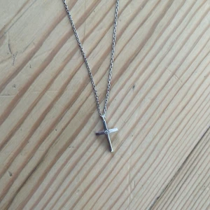Silverhalsband med korsberlock - Stilrent halsband i silverfärg med en tunn kedja och ett kors som hänge. Korset har en liten detalj i mitten som ger extra karaktär. Perfekt för dig som gillar minimalistiska smycken med en klassisk touch.