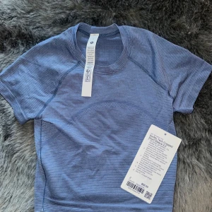 Blå croppad t-shirt från Lululemon - Superskön blå croppad t-shirt från Lululemon med diskreta ränder och rund hals. Fick den av mina föräldrar när de hade varit i usa, kunde tyvärr inte lämna tillbaks 🫶🏼 HELT OANVÄNDA!!T-shirten är kortärmad och har en tight passform, perfekt för träning.Materialet är mjukt och stretchigt, vilket gör den riktigt bekväm att bära.
