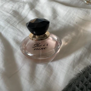 She is mine parfym med svart lock - Säljer en elegant parfymflaska med texten 'She is mine'. Flaskan är rund och transparent med en svagt rosa tonad vätska. Locket är stort, svart och diamantformat med en guldfärgad ring vid halsen. Perfekt för dig som gillar snygga detaljer på sminkbordet.