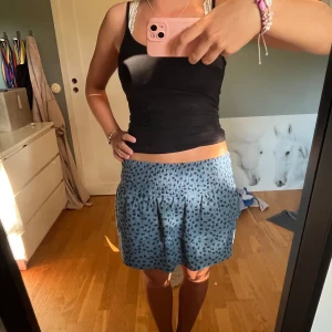 Volangkjol - Supersöt blå kortkjol med små svarta blommor och bred smock i midjan. Kjolen har en avslappnad passform och är perfekt för varma dagar. Tyget känns lätt och flowy, vilket gör den riktigt skön att bära. Sitter som en xs-s