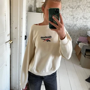Vit Reebok sweatshirt med broderad logo - Vit sweatshirt från Reebok med broderad logo i svart och rött framtill. Tröjan har en loose passform, rund halsringning och långa ärmar. Mjuk och skön i bomullsmix, perfekt till jeans eller leggings.