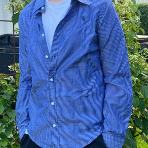 Blå randig skjorta från Polo Ralph Lauren - Säljer en klassisk blå skjorta från Polo Ralph Lauren med tunna vita ränder och broderad logga på bröstet. Skjortan har lång ärm, knappar framtill och är gjord i mjuk bomull. Perfekt för dig som gillar stilrena plag. Modellen är 162 och väger 45kg. Så jag skulle säga att den passar perfekt på 12-14  åringa killar. Storleken är 16 vilken passar för tjejer från 12-16.        Måtten är 57 i längd och 36 i axel längd.                  Skickas inom 1-2 dagar oftast!