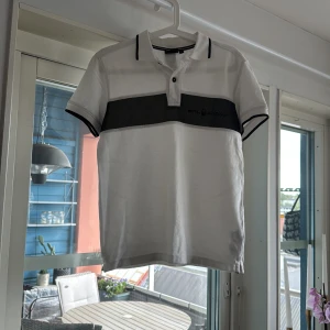 Vit polo t-shirt med svart detalj - Vit polo t-shirt från Still Racing med svart horisontell panel över bröstet och svarta detaljer på krage och ärmslut. Klassisk pikékrage med två knappar framtill. Perfekt för en clean och sportig look.