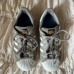 Adidas Superstar i grå mocka - Snygga Adidas Superstar sneakers i grå mocka med klassiska vita ränder och vit tå. Skorna har snörning och platt sula, samt detaljer i guld på sidan. Perfekta för en stilren och avslappnad look.